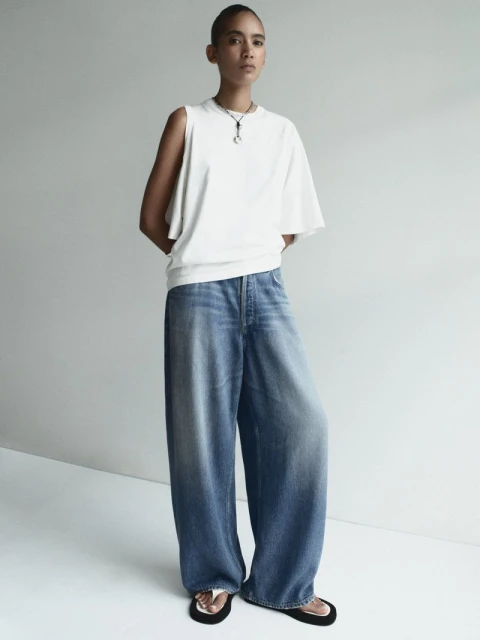 An adult woman poses indoors wearing a white, asymmetrical top, blue denim pants, and black slide sandals against a neutral background.