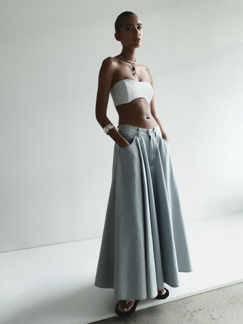 A fashion shot of an adult female model in a strapless top and denim skirt against a light grey studio backdrop, showcasing fashion and style.
