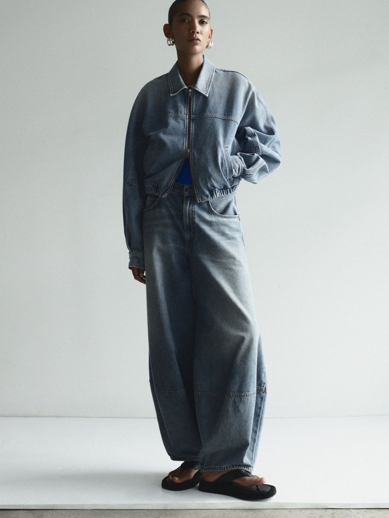 A young adult posing in light-wash denim jacket and wide-leg jeans, complemented by dark-colored sandals against a neutral backdrop.