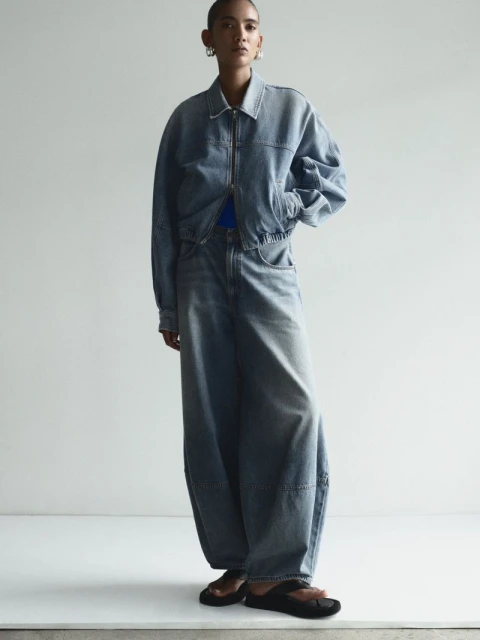 A young adult posing in light-wash denim jacket and wide-leg jeans, complemented by dark-colored sandals against a neutral backdrop.