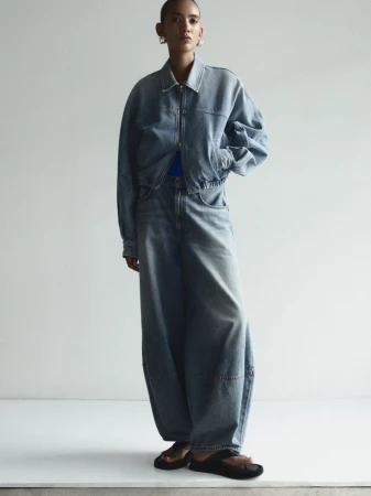 A young adult posing in light-wash denim jacket and wide-leg jeans, complemented by dark-colored sandals against a neutral backdrop.