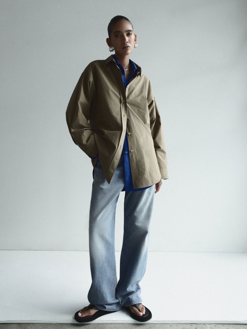 Woman in oversized khaki shirt and jeans posing in front of a white wall in a fashion editorial, wearing silver hoop earrings.