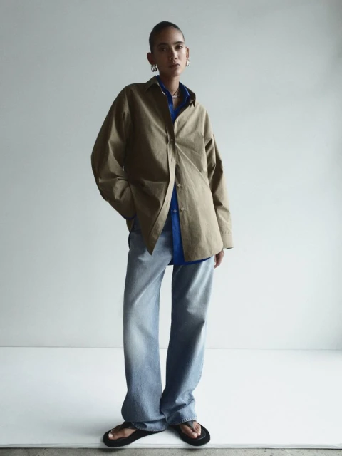 Woman in oversized khaki shirt and jeans posing in front of a white wall in a fashion editorial, wearing silver hoop earrings.