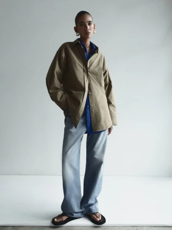 Woman in oversized khaki shirt and jeans posing in front of a white wall in a fashion editorial, wearing silver hoop earrings.
