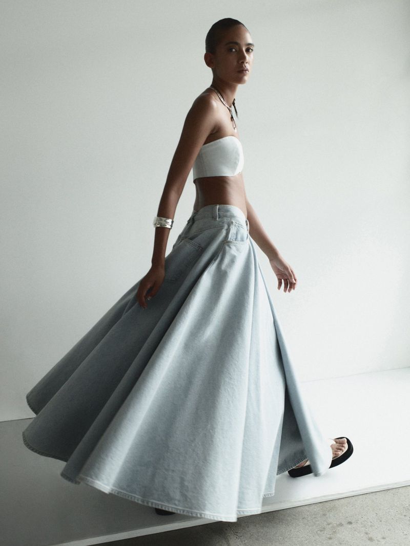 Fashionable woman strides in a white tube top and flowing light blue denim skirt in a studio. Minimalist studio fashion shot.