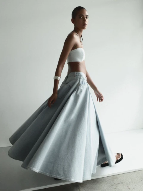 Fashionable woman strides in a white tube top and flowing light blue denim skirt in a studio. Minimalist studio fashion shot.