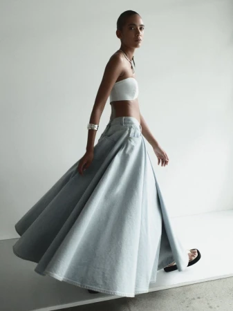 Fashionable woman strides in a white tube top and flowing light blue denim skirt in a studio. Minimalist studio fashion shot.