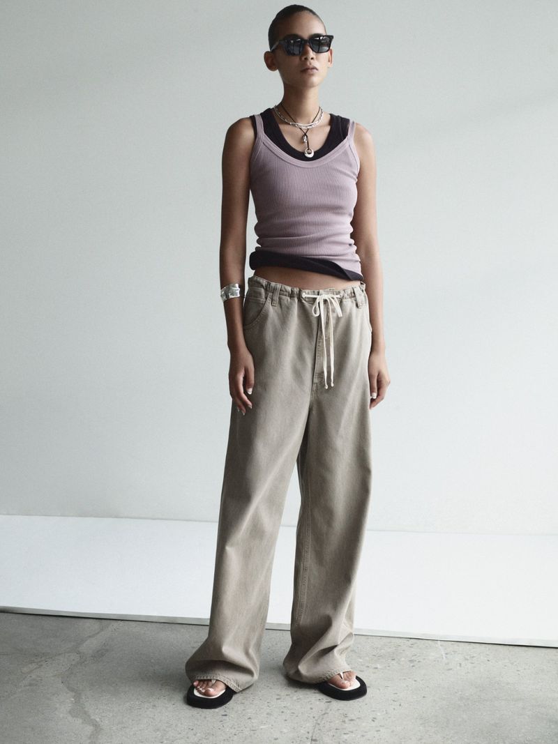 Young woman in a lilac ribbed tank top and khaki wide-leg pants stands in a minimalist setting with neutral background, wearing sunglasses.