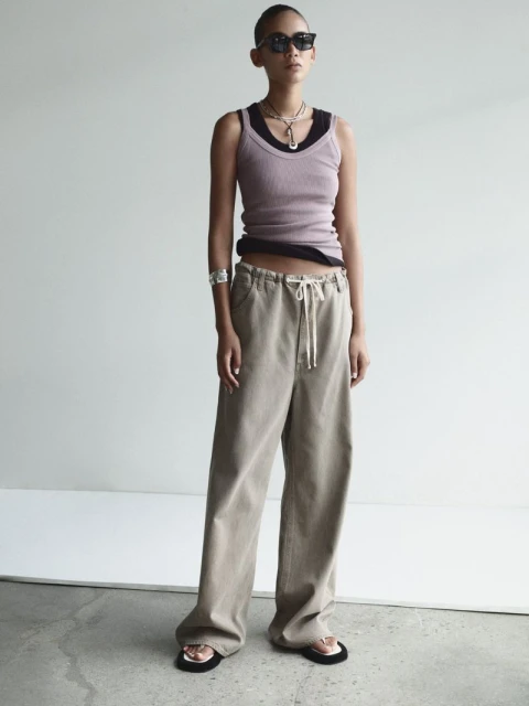 Young woman in a lilac ribbed tank top and khaki wide-leg pants stands in a minimalist setting with neutral background, wearing sunglasses.