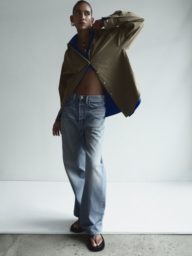 Fashion shot of a young adult woman posing indoors wearing a brown jacket, blue jeans, and black flip-flops in a modern editorial style.