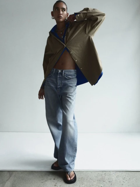 Fashion shot of a young adult woman posing indoors wearing a brown jacket, blue jeans, and black flip-flops in a modern editorial style.
