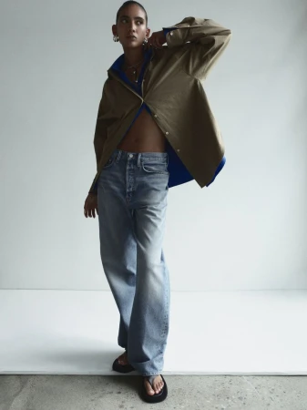 Fashion shot of a young adult woman posing indoors wearing a brown jacket, blue jeans, and black flip-flops in a modern editorial style.
