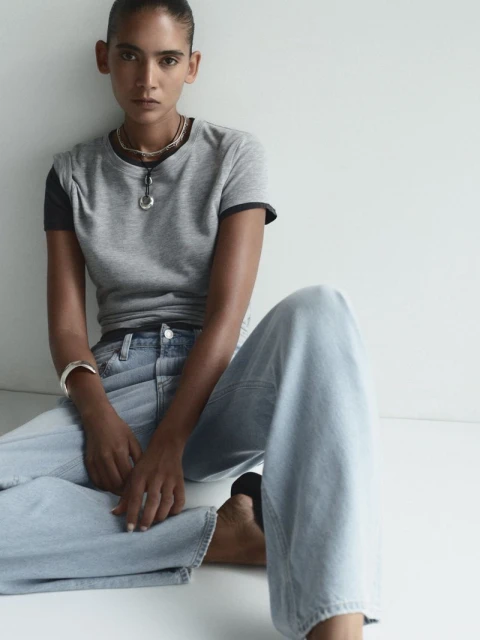 A young adult posing indoors, wearing a grey T-shirt and jeans. Minimalist fashion shot with soft light and a neutral studio backdrop.