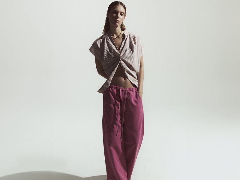 A female fashion model poses in a striped, short-sleeve blouse and magenta pants in a studio with soft lighting and subtle shadows.