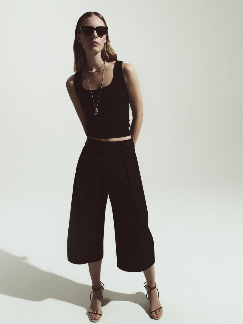 Full-length shot of a woman in a black tank top and culottes, accessorized with sunglasses and a necklace, posing in a studio setting.
