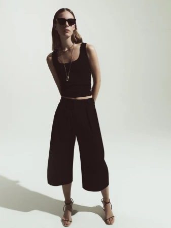 Full-length shot of a woman in a black tank top and culottes, accessorized with sunglasses and a necklace, posing in a studio setting.