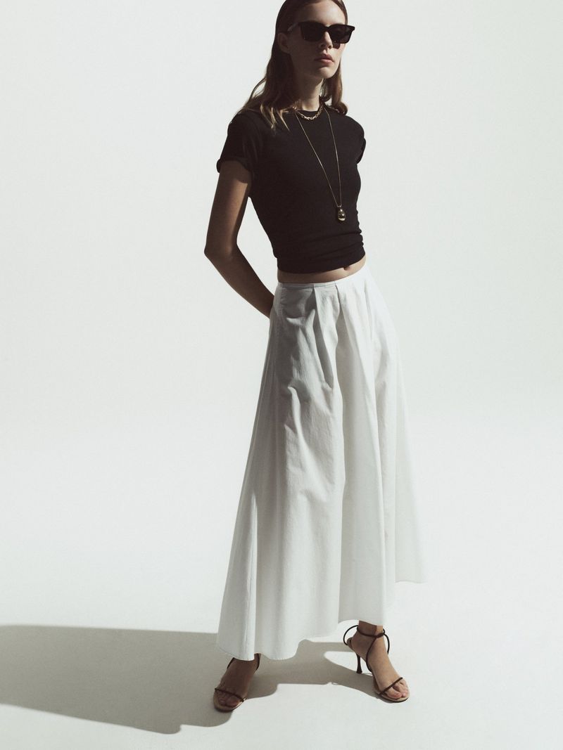 A slender adult woman poses in a black crop top and long white skirt against a bright white background, styled with black sunglasses.