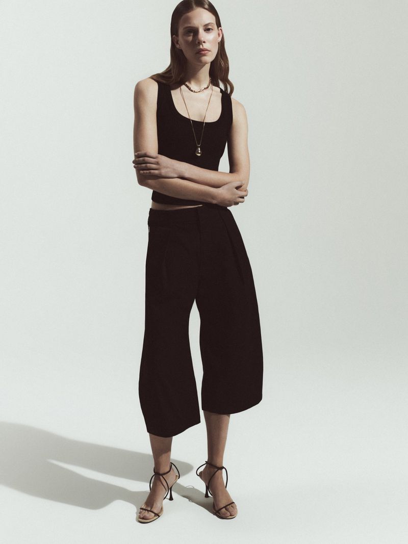 Studio shot of a woman wearing a black tank top, culottes, and strappy sandals with jewelry, posing in a neutral setting.