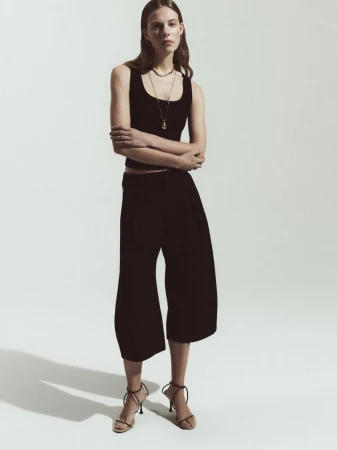 Model Anouk Smits standing in a black sleeveless top, black cropped culottes, and strappy heels against a white studio background.