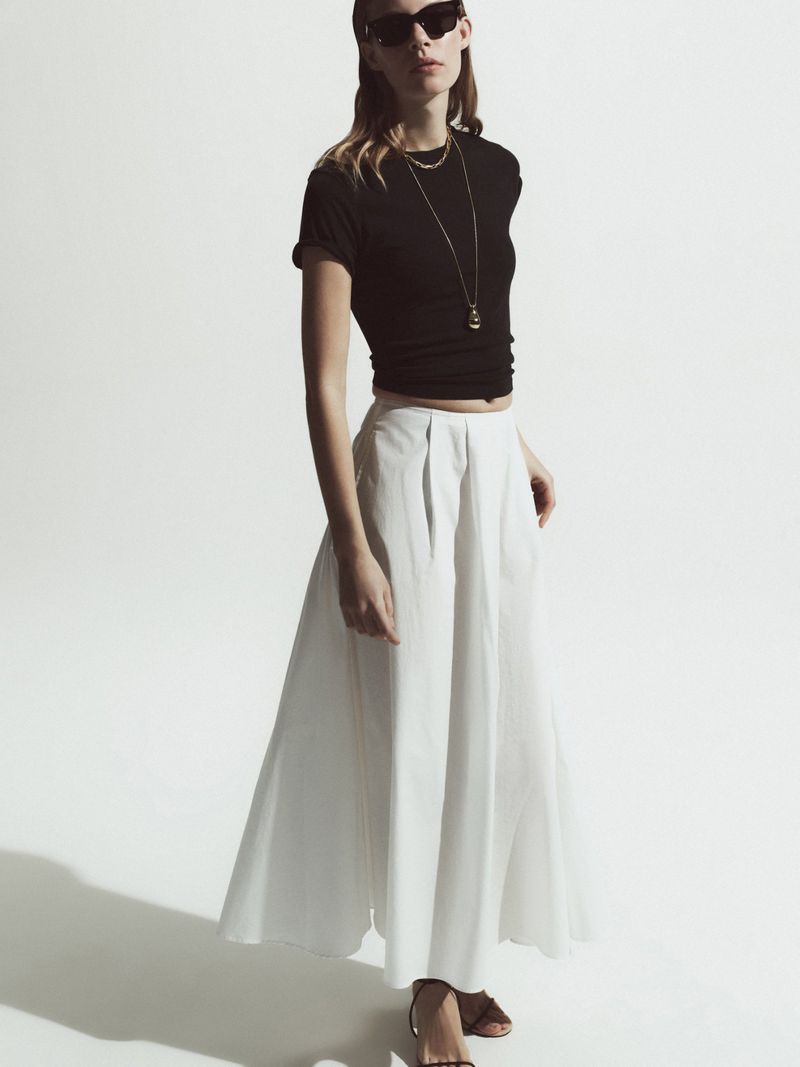 A woman poses in a black t-shirt and a white pleated skirt, accessorized with necklaces and sunglasses, against a plain studio backdrop.