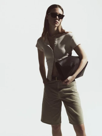 Woman in button-down shirt, Bermuda shorts, and sunglasses holding a bag against a white backdrop in a fashion editorial.