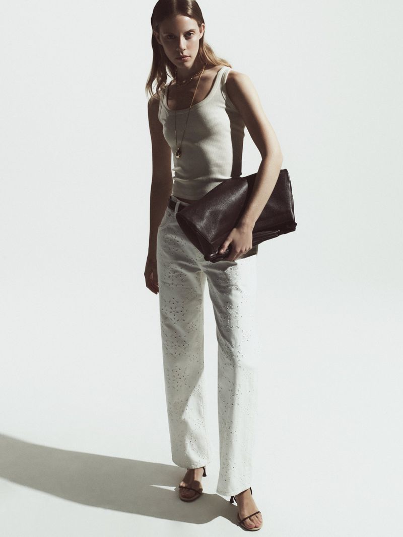 Fashionable adult female poses in a studio wearing a tank top and white trousers with strappy heeled sandals, holding a clutch purse.