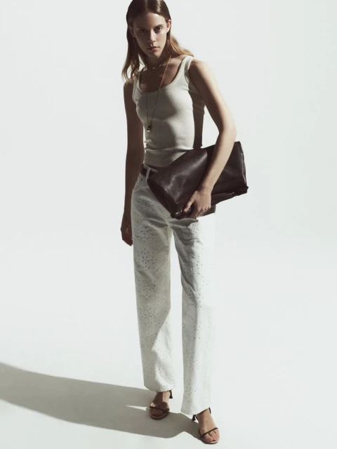 Fashionable adult female poses in a studio wearing a tank top and white trousers with strappy heeled sandals, holding a clutch purse.