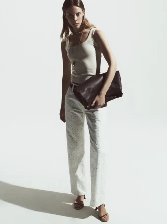 Fashionable adult female poses in a studio wearing a tank top and white trousers with strappy heeled sandals, holding a clutch purse.