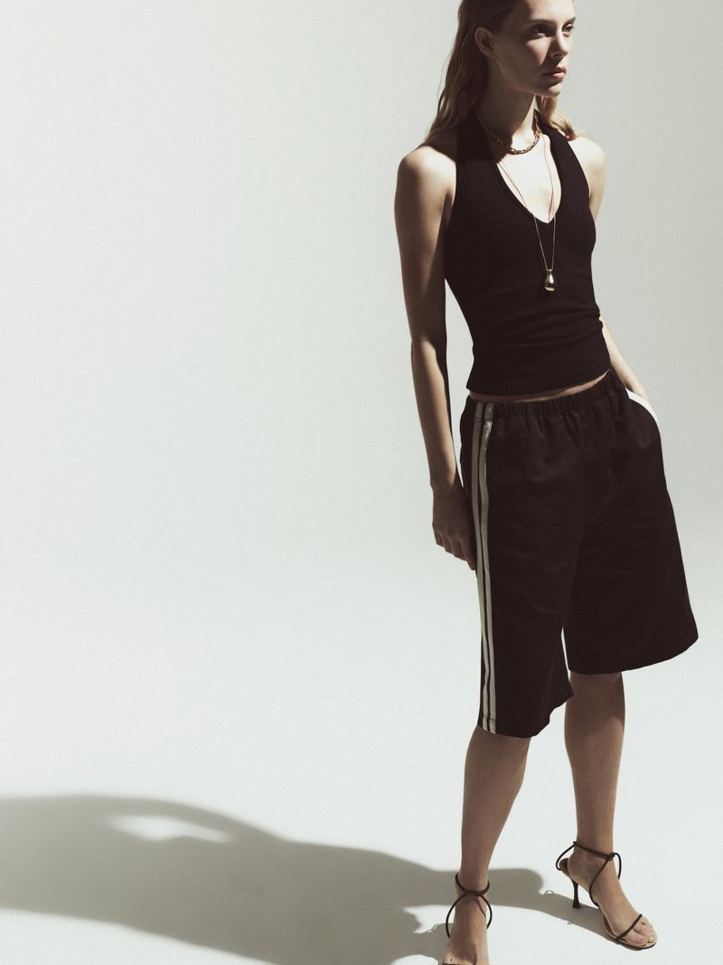 A woman poses in a black halter top, black shorts with white stripes, and high-heeled sandals against a bright white background.