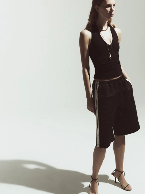 A woman poses in a black halter top, black shorts with white stripes, and high-heeled sandals against a bright white background.