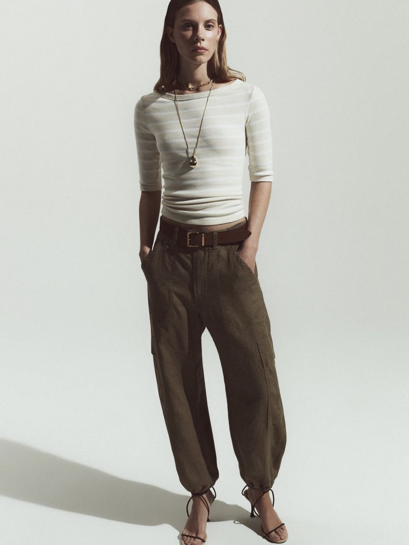 Full-length shot of a woman in a striped top and baggy green pants with strappy heels against a bright white seamless backdrop.