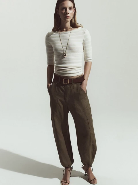 Full-length shot of a woman in a striped top and baggy green pants with strappy heels against a bright white seamless backdrop.