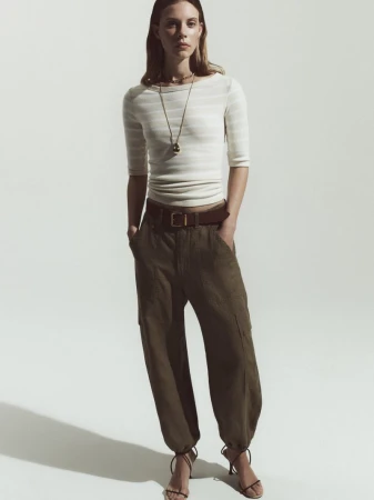Full-length shot of a woman in a striped top and baggy green pants with strappy heels against a bright white seamless backdrop.