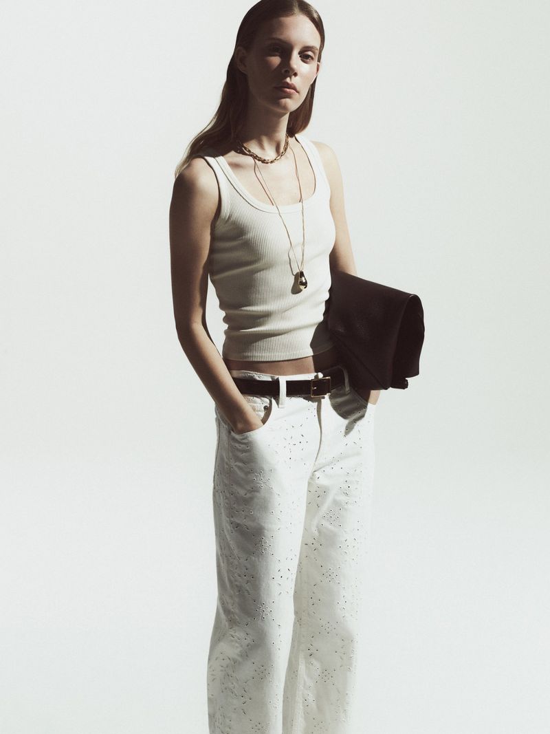 A stylish young woman in a cream tank top and white jeans stands indoors holding a black jacket over her shoulder.