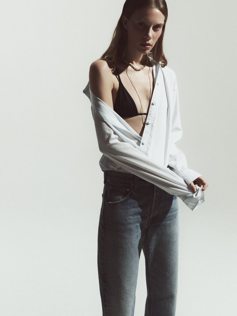Fashion shot of a woman wearing a black bikini top, blue jeans, and a white blouse, posing against a bright white backdrop.