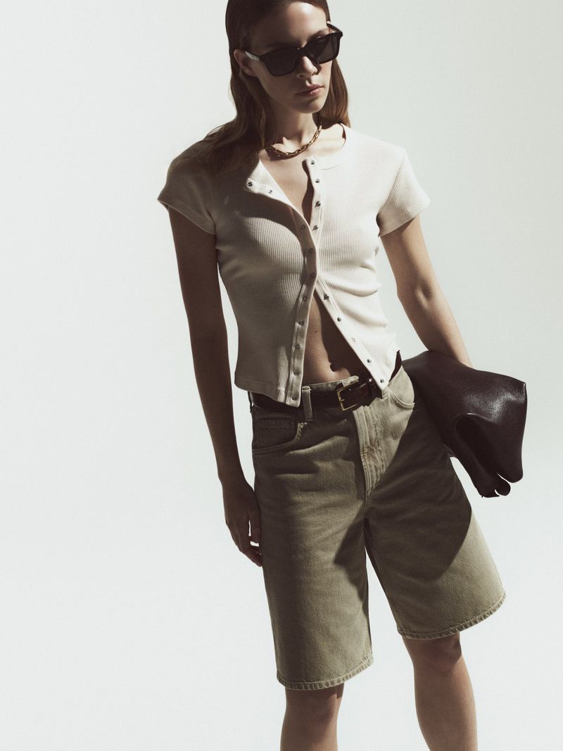 A woman wearing a buttoned shirt, olive shorts, and sunglasses stands in front of a neutral background, holding a dark purse.