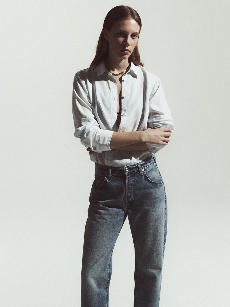 Portrait of an adult woman posing with arms crossed, wearing a white button-down shirt and denim blue jeans, standing indoors on a white background.