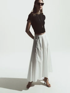 Model Anouk Smits posing in dark sunglasses, a fitted black t-shirt, and a white maxi skirt against a bright white studio backdrop.