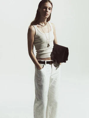 Model Anouk Smits wears a white ribbed tank top and white eyelet trousers with a dark belt against a bright white background.