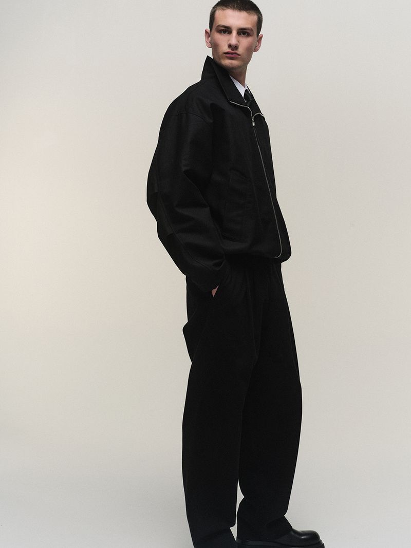 A man standing in profile wearing a black zip jacket, matching black trousers, and a white collared shirt against a pale studio backdrop.