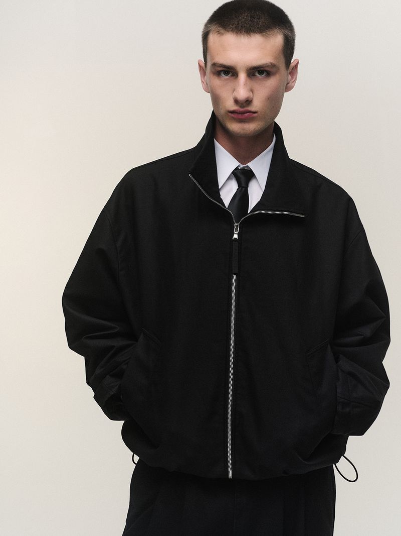 A man wearing an oversized black zip-up jacket, white collared shirt, and black tie posing against a pale seamless studio backdrop.