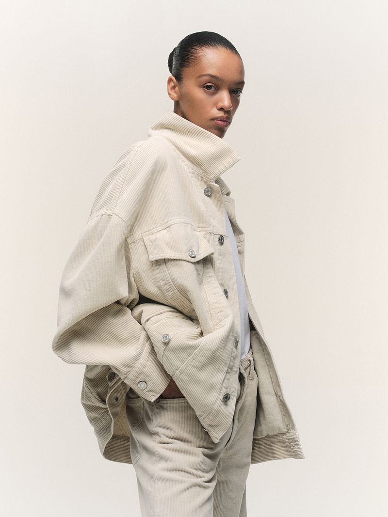 Close-up of a young adult woman posing in a corduroy jacket and pants, set against a neutral backdrop in a fashion studio setting.