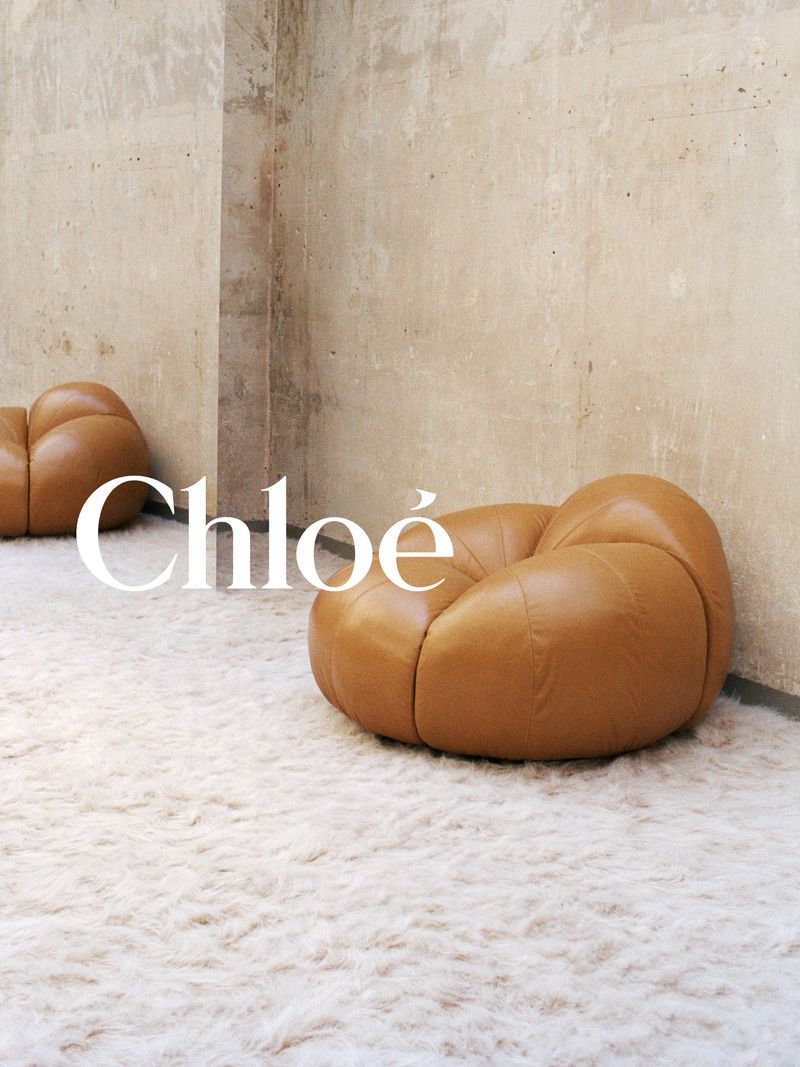 Two tan leather pouf chairs sit on a white shag rug against a tan plaster wall, illuminated by soft, diffused light, with Chloé logo.