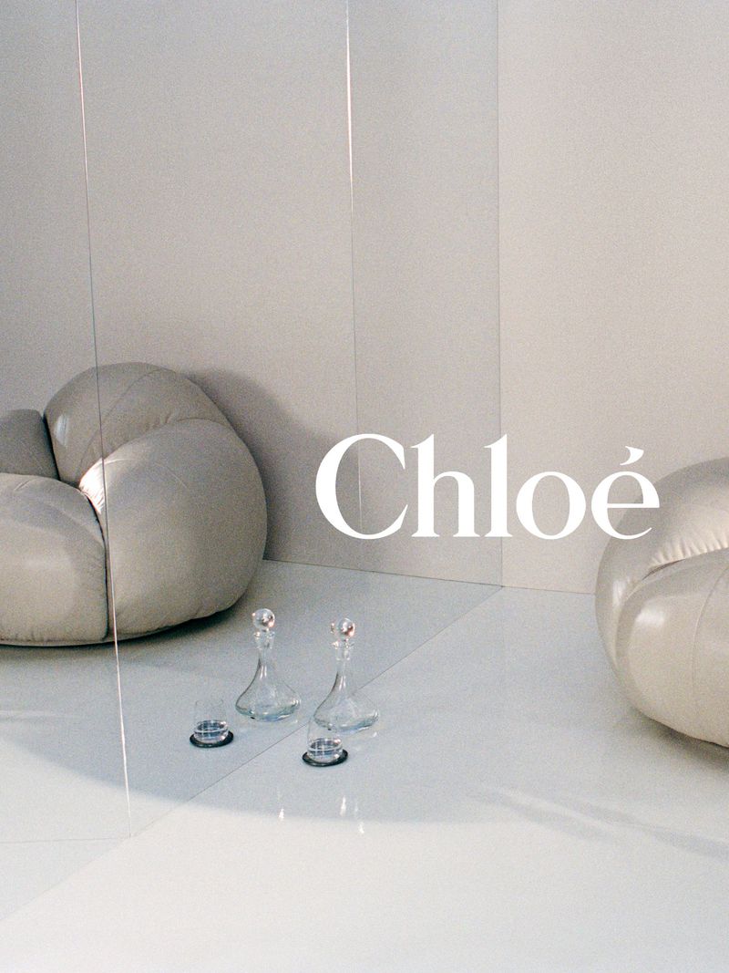 Minimalist composition featuring a Chloe logo, two glass decanters, and a rounded sofa in a bright, neutral-toned interior setting.