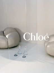 Minimalist interior set featuring sculptural cream armchairs, glass decanters, and the Chloé logo on glossy white flooring.