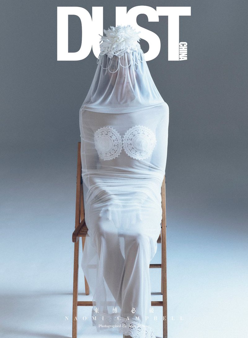 Fashion magazine cover featuring a model in a white lace bra and sheer fabric, seated on a wooden chair against a grey background.