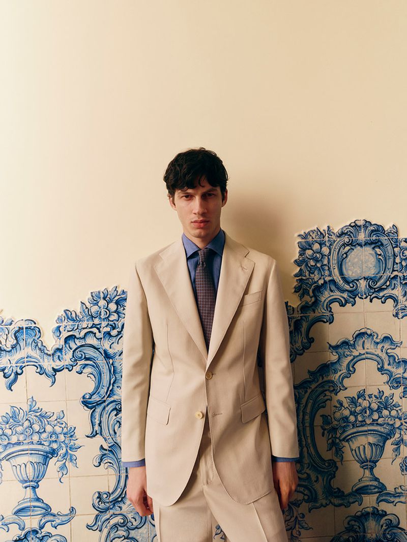 Portrait of a dark-haired man in a tan suit, blue shirt, and patterned tie, posed in front of a decorative tile wall.