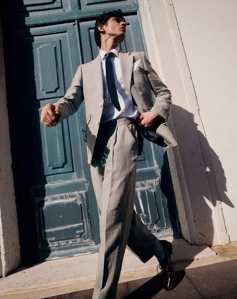 A stylish man in a grey suit walks confidently past a teal door in bright sunlight, accessorized with a tie and pocket square.