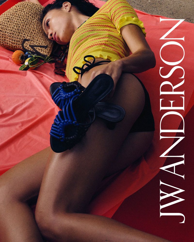 Woman in a yellow top reclines on a red sheet with blue sandals, a straw bag, and fruit beside her, featuring the JW Anderson logo.