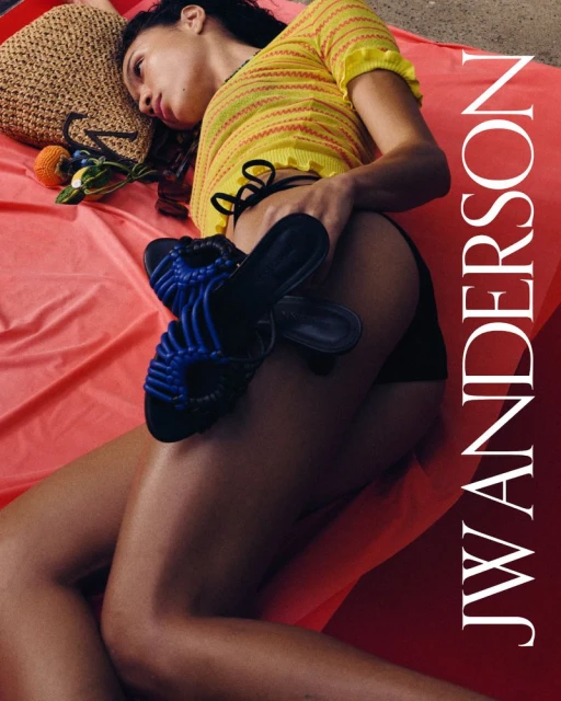 Woman in a yellow top reclines on a red sheet with blue sandals, a straw bag, and fruit beside her, featuring the JW Anderson logo.